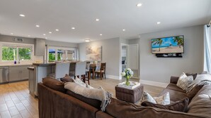 A modern living room with a brown sofa, a dining area with a table and chairs, a kitchen with grey cabinets, and a TV mounted on the wall displaying a beach scene. - SPECIALS!! BEAUTIFUL BEACH HOUSE ! WALK 2 BEACH POOL ! HOT TUB (Lauderdale-By-The-Sea)