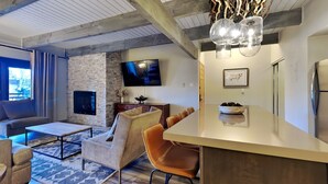 A modern living room with a stone wall, a flat-screen TV, a dining area with a bar, and a large window. - 🌲Timberline D2B Premier 1-Bedroom ~Hot Tub~Pool~WiFi~Ski-in/out~Local Shuttle🌲 (Snowmass Village)