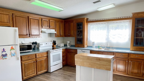 Una cocina con armarios de madera, un refrigerador blanco, un microondas, un horno, un lavavajillas y una ventana con vistas.