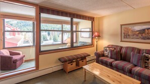 A cozy living room with a large window, a sofa, a bench, and a lamp. - Spacious condo just steps away from main lifts, hot tub, free wifi & parking (Copper Mountain)