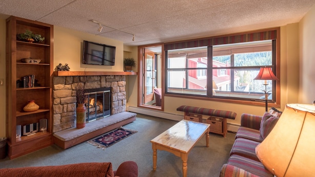 A cozy living room with a stone fireplace, a television mounted on the wall, a large window with a view of buildings, and a comfortable seating area with a sofa and a bench. - Spacious condo just steps away from main lifts, hot tub, free wifi & parking (Copper Mountain)