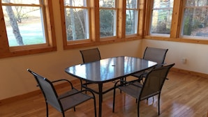 A dining area with a glass-top table and four chairs, large windows with wooden frames, and wooden flooring.