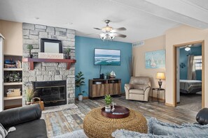 Ein Wohnzimmer mit Kamin aus Stein, Deckenventilator, Wand-Fernseher und Meerblick durch ein Fenster.
