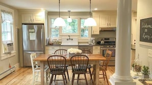 Una cocina moderna con armarios blancos, un refrigerador de acero inoxidable, una mesa de comedor de madera con sillas, lámparas colgantes y un letrero de pizarra.
