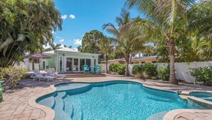 A swimming pool with a tiled deck, surrounded by palm trees and a building with large windows. - Mermaid Cove: Updated Forida Cottage w/pool, hot tub (Anna Maria)