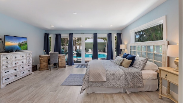 A bedroom with a bed, dresser, TV, and a view of a pool and outdoor area. - Mermaid Cove: Updated Forida Cottage w/pool, hot tub (Anna Maria)