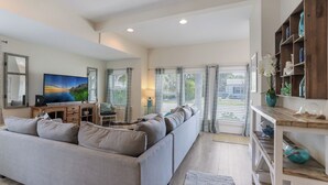 A modern living room with a grey sectional sofa, a flat-screen TV, a wooden coffee table, and a bookshelf. - Mermaid Cove: Updated Forida Cottage w/pool, hot tub (Anna Maria)