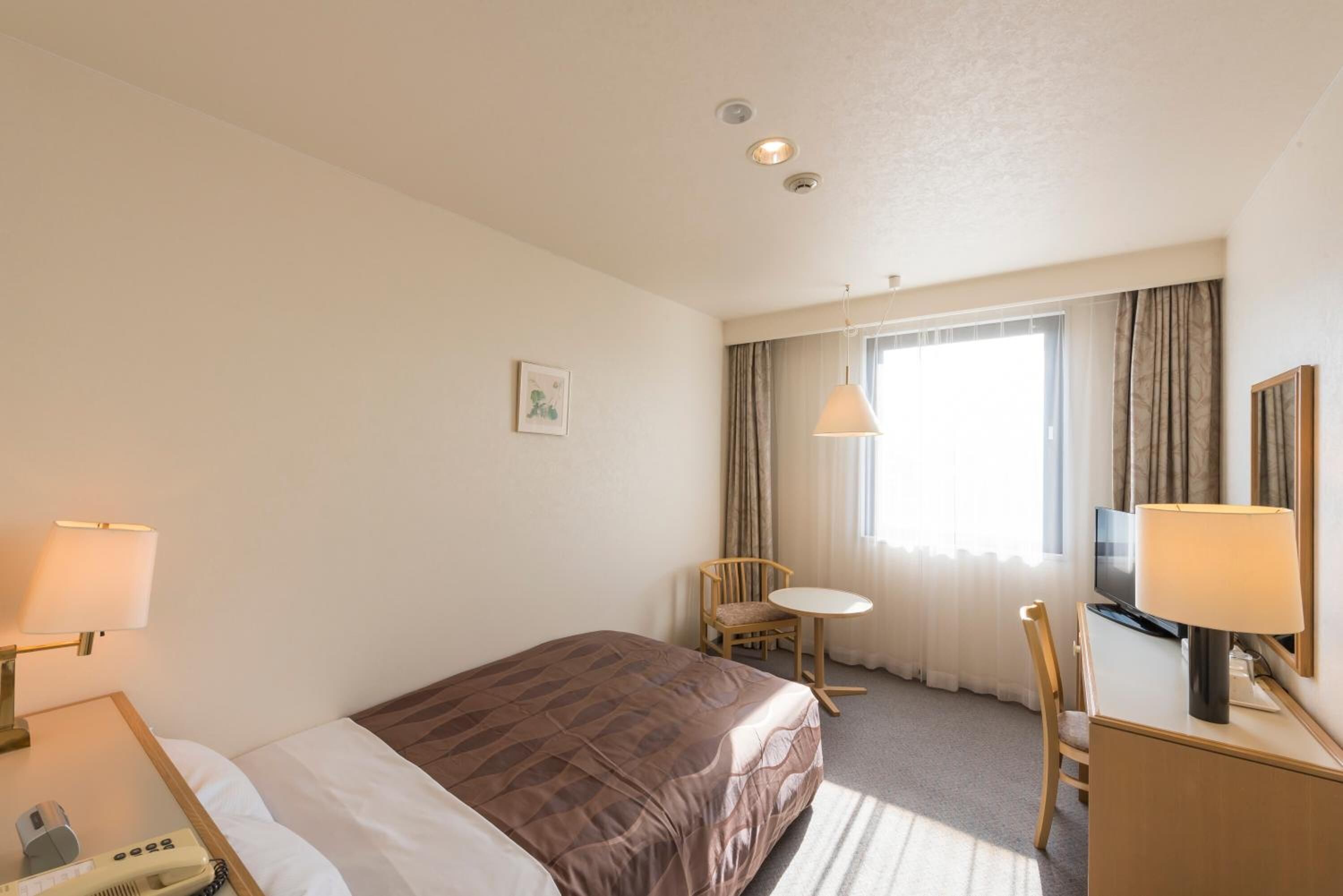 A hotel room with a bed, bedside table, lamp, chair, desk, TV, and a window with curtains.