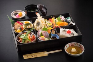 Una caja bento japonesa tradicional con diferentes tipos de sushi y acompañamientos, junto con palillos y un tazón pequeño de sopa.