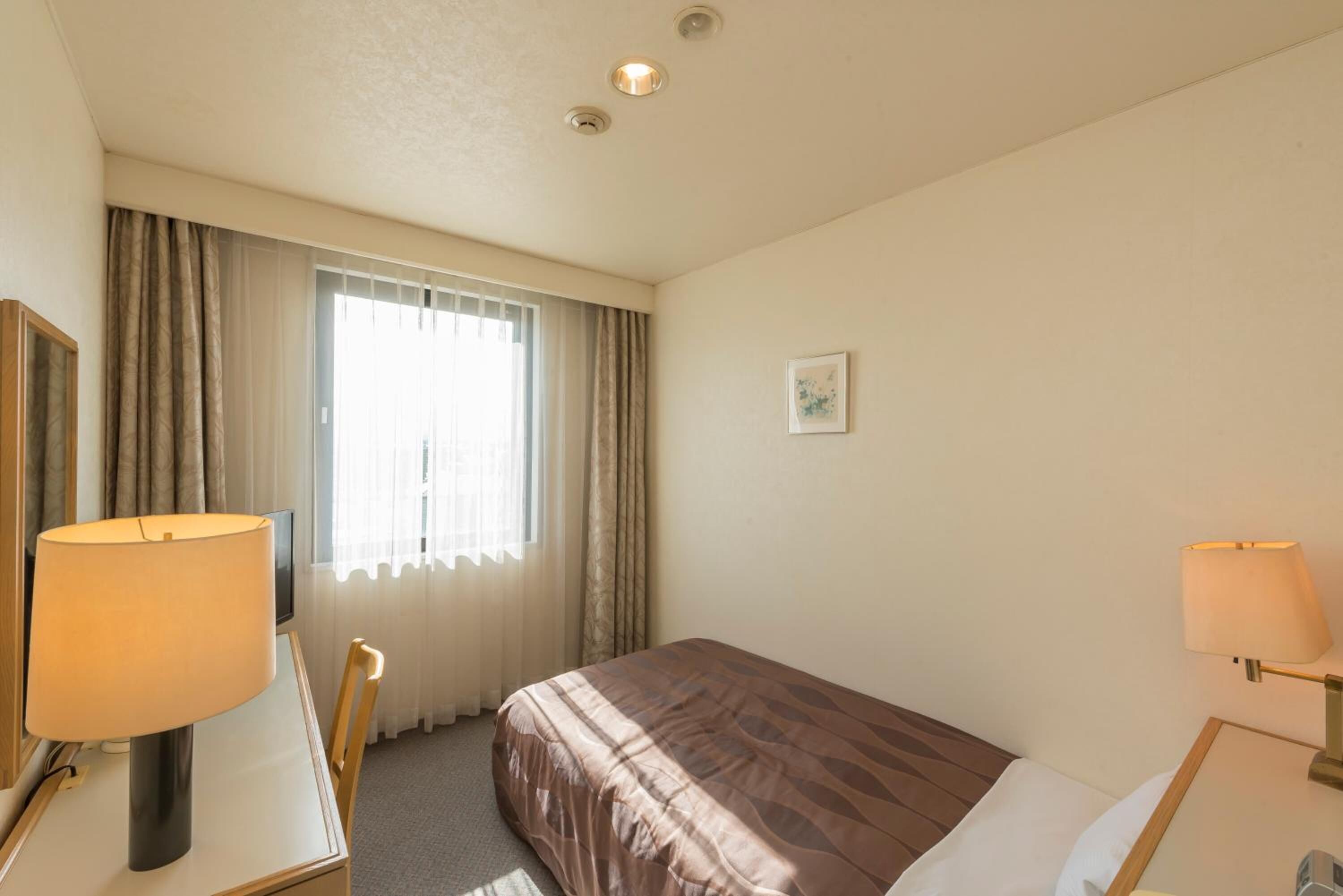 Una habitación de hotel con una cama, un escritorio con lámpara, una silla, un ventanal con cortinas y un cuadro pequeño en la pared.