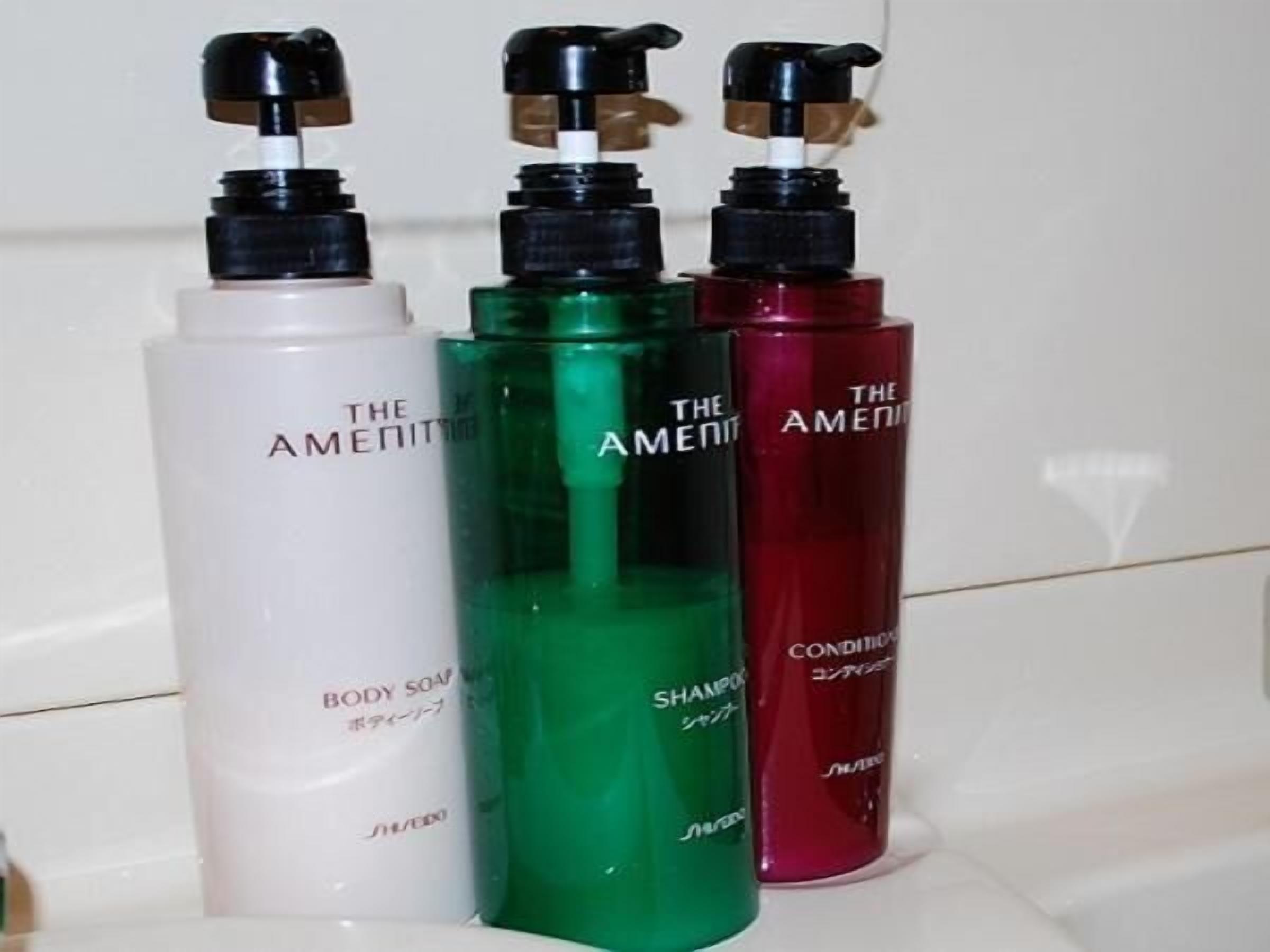 Trois flacons du savon pour le corps, du shampooing et de l’après-shampooing de la marque The Amenity, posés sur une surface blanche.