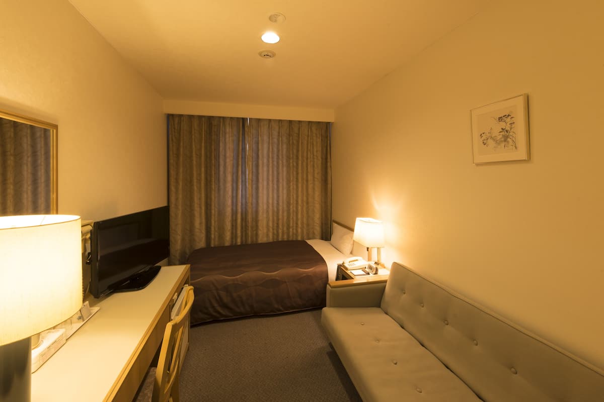 Una habitación de hotel con cama, escritorio con lámpara, televisor, mesita de noche con lámpara y un cuadro en la pared.