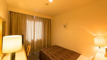 A hotel room with a single bed, a desk with a lamp, a chair, and a window with curtains.