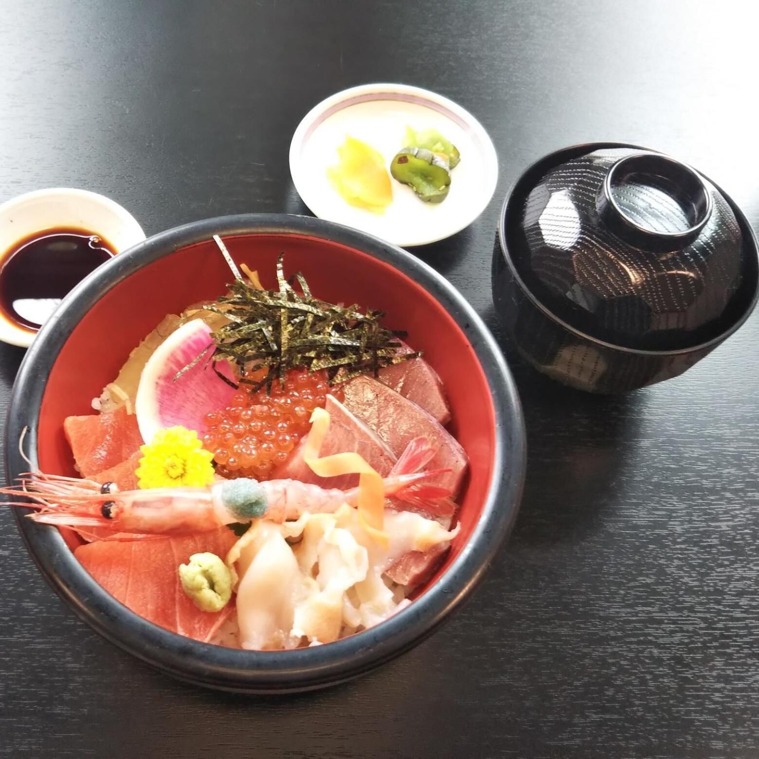 A bowl of sashimi with various seafood, including shrimp, fish, and roe, accompanied by small dishes of soy sauce and wasabi.