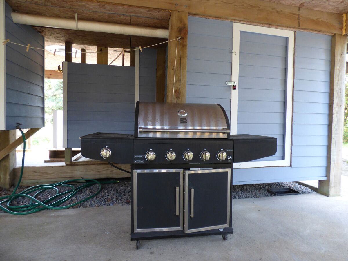 A barbecue grill with a built-in oven and a side cabinet, set up on a concrete patio under a wooden structure.