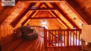 A wooden attic room with a wooden ceiling, a chair, a table, and a railing.