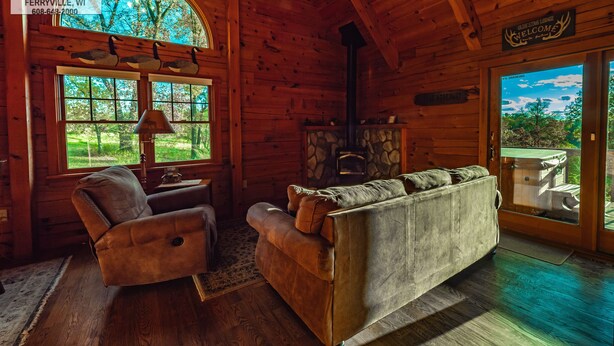 A cozy cabin interior with wooden walls, a fireplace, a sofa, and a dining area with a view of the outdoors.
