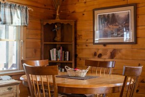 Uma mesa de jantar de madeira com quatro cadeiras, uma cesta trançada sobre a mesa e uma estante de livros ao fundo.