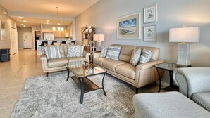 A spacious living room with a beige sofa set, a glass coffee table, and a grey rug. There is a kitchen in the background with modern appliances and a dining area.