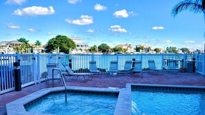 A pool area with lounge chairs and a small hot tub, surrounded by a white fence, with a view of buildings in the background.
