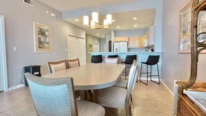 A spacious dining area with a large round table and six chairs, a kitchen in the background, and a chandelier.