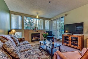 A living room with a sofa, a coffee table, a fireplace, a TV, and large windows with blinds.