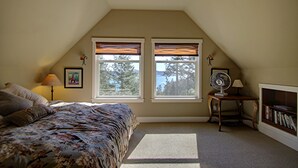 Un dormitorio con una cama, dos ventanas con persianas, un ventilador, una lámpara y una estantería.
