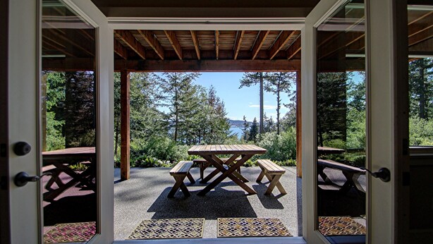 Un patio abierto con una mesa y bancos de madera, rodeado por una pérgola de madera, con vistas a árboles y una masa de agua al fondo.
