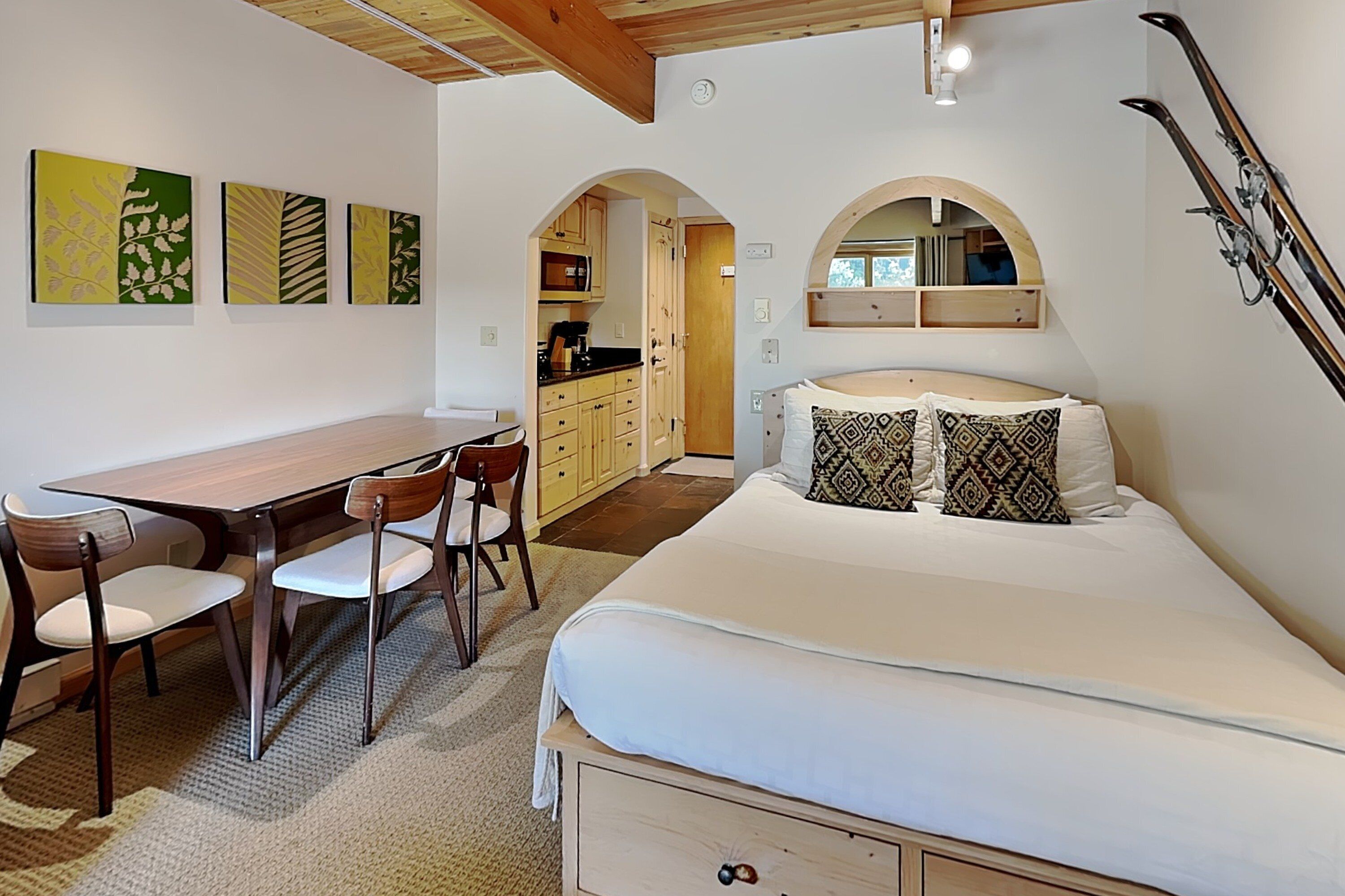 A bedroom with a bed, a desk, chairs, and a kitchen area.