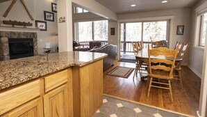 A spacious kitchen with granite countertops, wooden cabinets, and a stone fireplace. There is a dining area with a wooden table and chairs, and large windows offering views of the outdoors. - Spacious Town Home with Ski-In/Ski-Out Access (Whitefish)