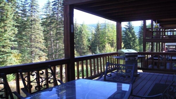 A wooden deck with a glass table and chairs, overlooking a forested area. - Spacious Town Home with Ski-In/Ski-Out Access (Whitefish)
