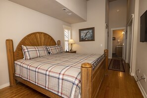 A bedroom with a wooden bed, a plaid bedspread, a nightstand with a lamp, and a framed picture on the wall.