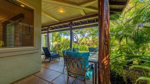A covered patio area with a table and chairs, surrounded by lush greenery and a view of the outdoors. - Tropical Villa w/Beach Club Access ★Quiet Location★Bikes and Beach Gear★Walk to Restaurants (Kamuela)