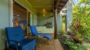 A patio with two blue chairs and a small table, a kitchen area with a grill, and a clock on the wall. - Tropical Villa w/Beach Club Access ★Quiet Location★Bikes and Beach Gear★Walk to Restaurants (Kamuela)