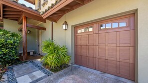 A residential garage with a brown door, a stone pathway, and a small garden with a palm plant. - Tropical Villa w/Beach Club Access ★Quiet Location★Bikes and Beach Gear★Walk to Restaurants (Kamuela)