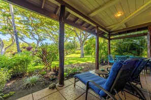 A covered outdoor seating area with a bench and chairs, surrounded by lush greenery and trees.