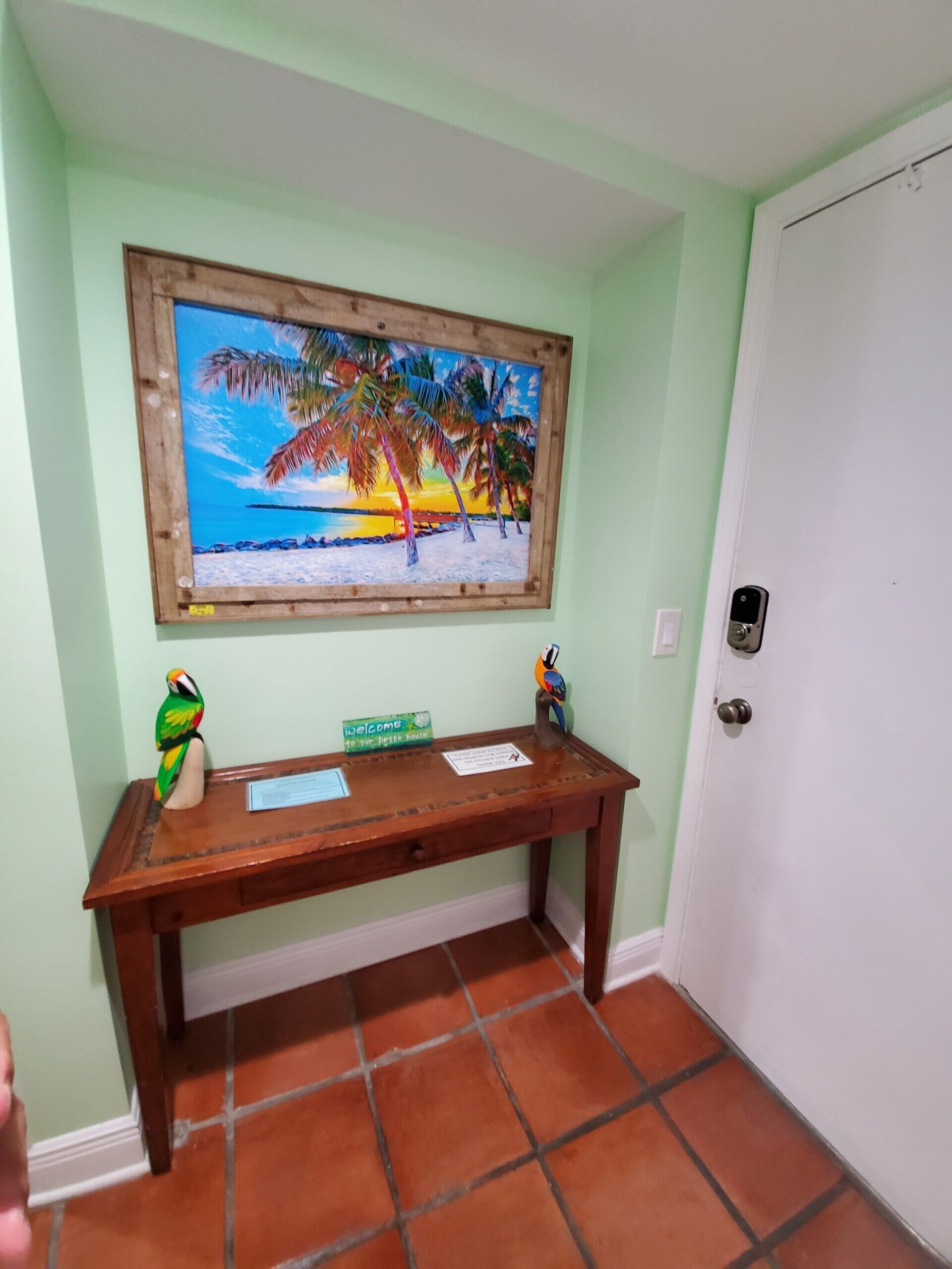 Un banco de madera con un cartel de bienvenida, un cuadro enmarco con una playa y una figura de un loro.