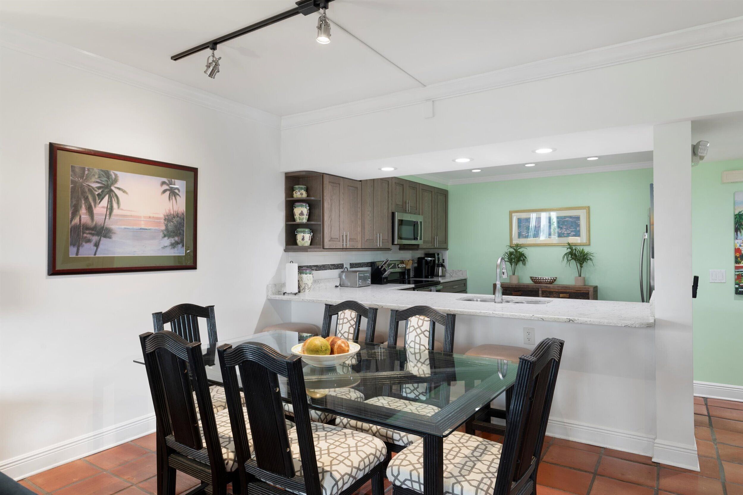 A dining area with a glass table and chairs, a kitchen with wooden cabinets, and a green accent wall.