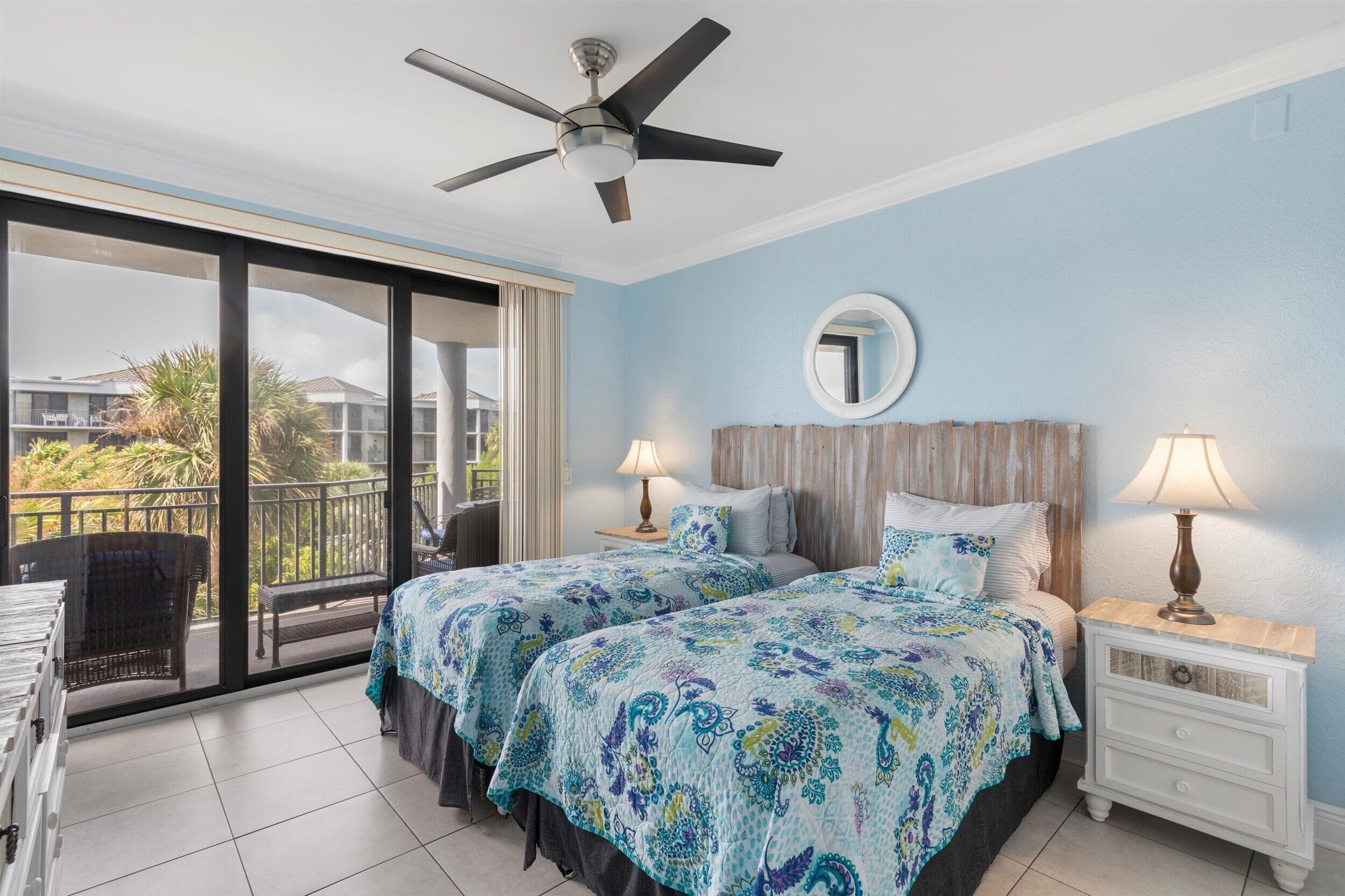 A hotel room with two beds, a balcony, a ceiling fan, and a view of the outside.