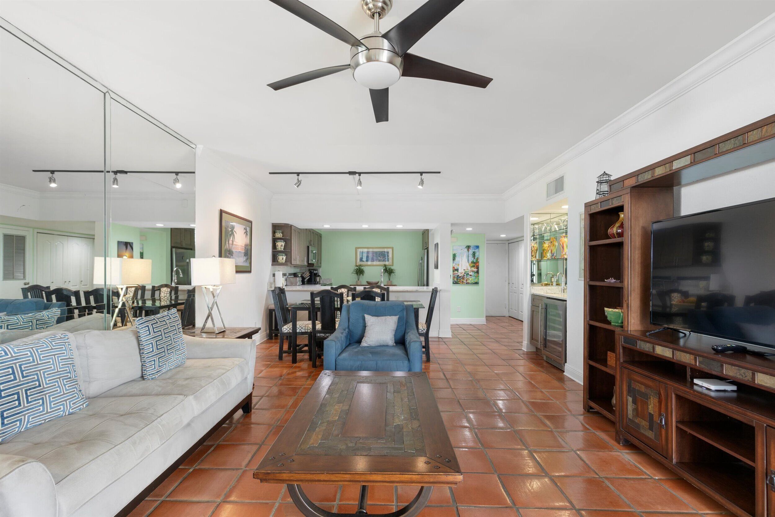 A spacious living room with a sofa, coffee table, and TV stand. A ceiling fan and kitchen are visible in the background.