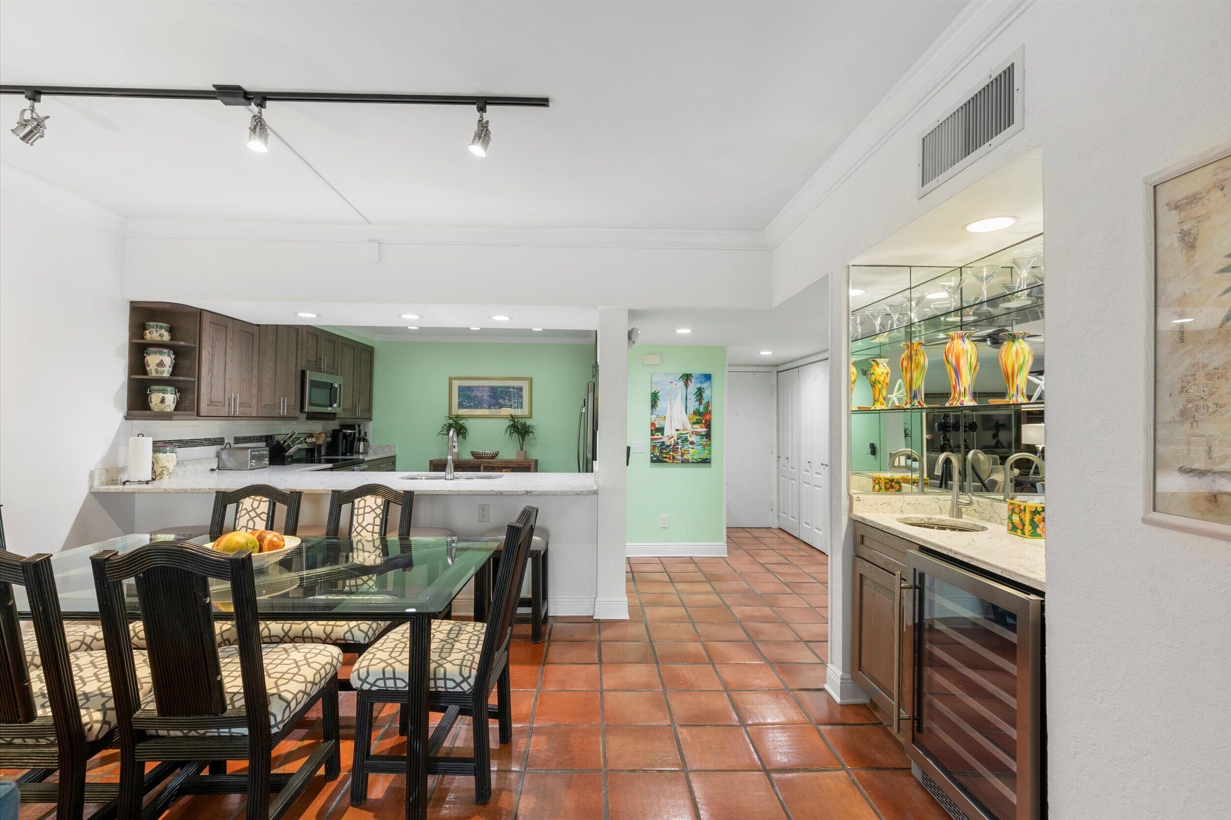 A modern kitchen with a dining area, featuring a glass table, wooden chairs, and a tiled floor.