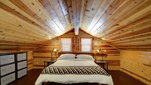 Un dormitorio de cabaña de madera con techo inclinado, dos mesitas de noche con lámparas, una cama con colcha estampada y un mueble de almacenamiento con varios cajones.