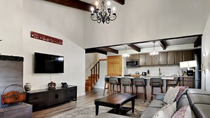Un salón espacioso con un sofá gris grande, una mesa de centro y un televisor montado en la pared. Cuenta con una zona de cocina con microondas y un comedor con sillas.