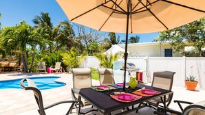 Une terrasse avec une table dressée pour le repas, entourée de chaises, d’une piscine et de palmiers.