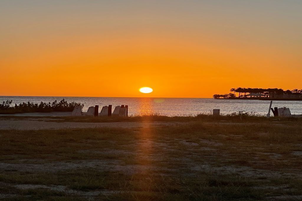 Un coucher de soleil sur une plage, avec un ciel dégagé, une mer calme et une côte lointaine parsemée de bâtiments.