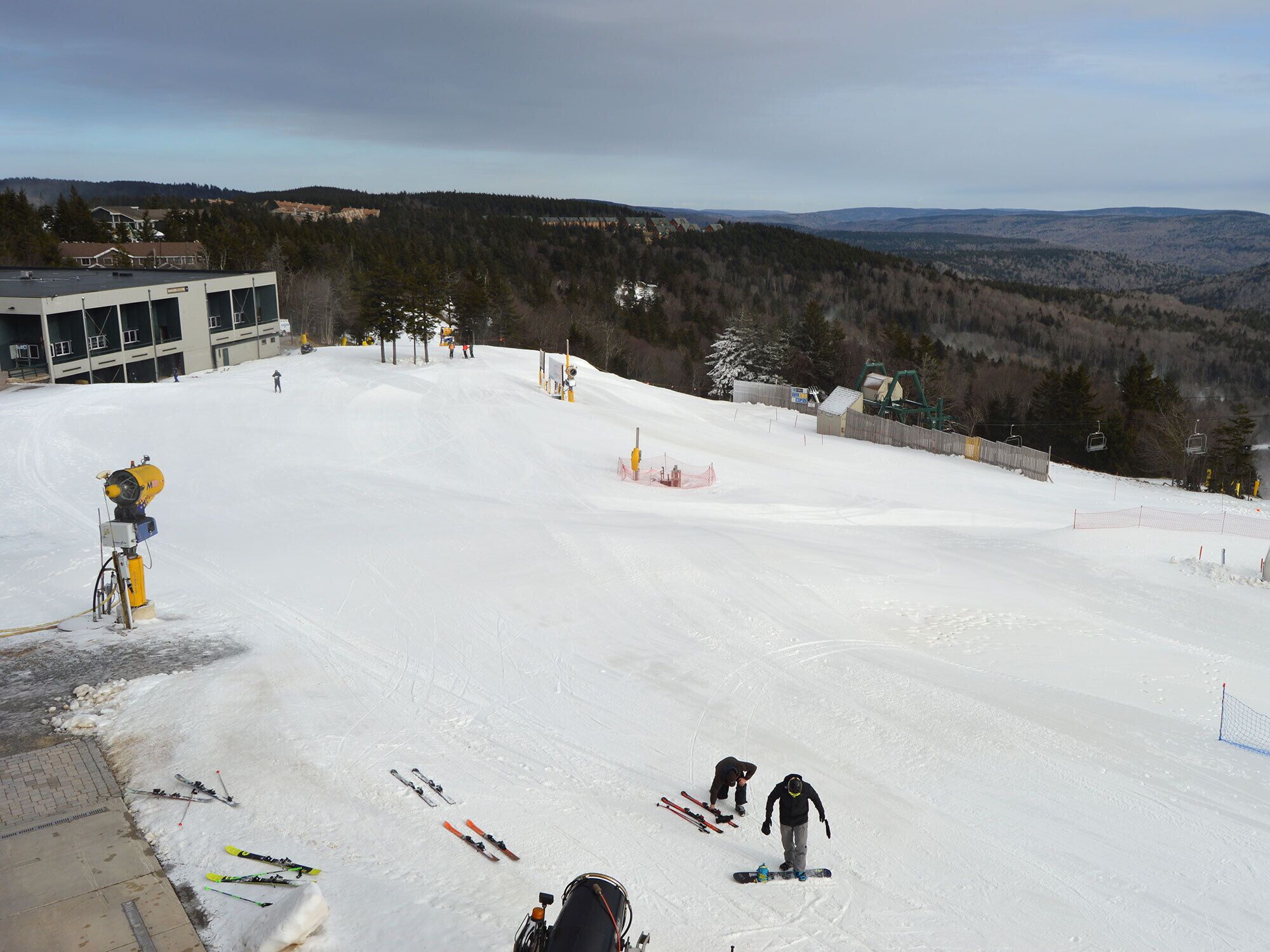 A ski slope with a building in the background, a snow-making machine, and a fence.