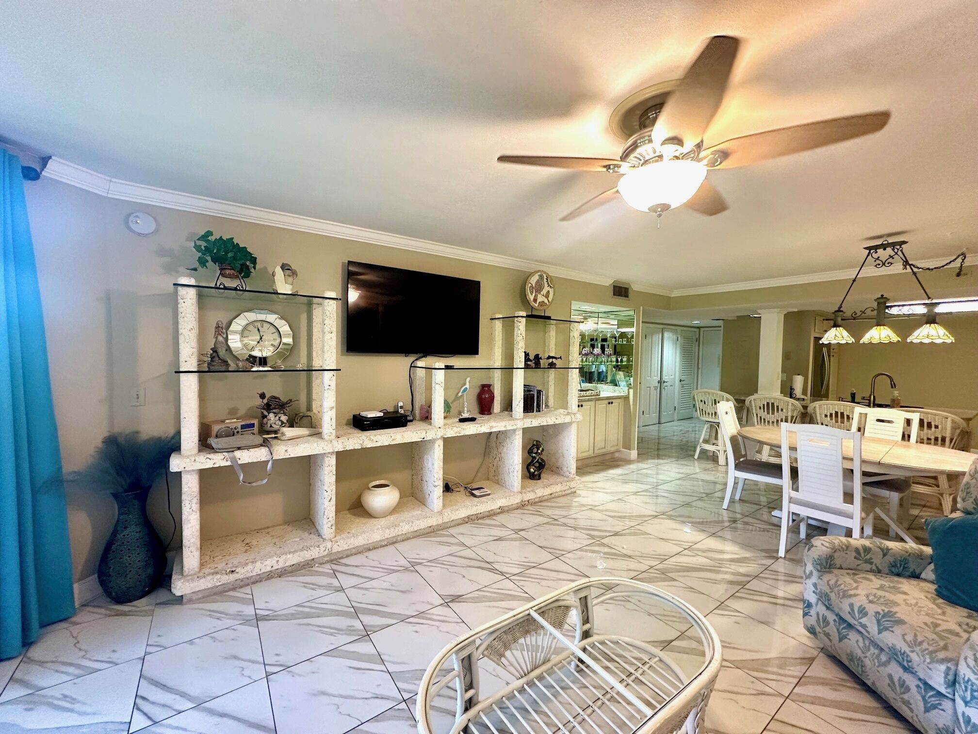 A living room with a TV, shelves with decorative items, a dining area with chairs and a table, and a ceiling fan.