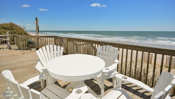 Terraza de madera con una mesa redonda y sillas blancas, con vista a una playa y olas.