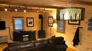 A cozy cabin interior with a leather sofa, a round table with a wood slice centerpiece, a fireplace, and framed pictures on the walls. - The Retreat - Cozy studio cabin - Hot Tub, Firepit, pond, natural playground (Logan)
