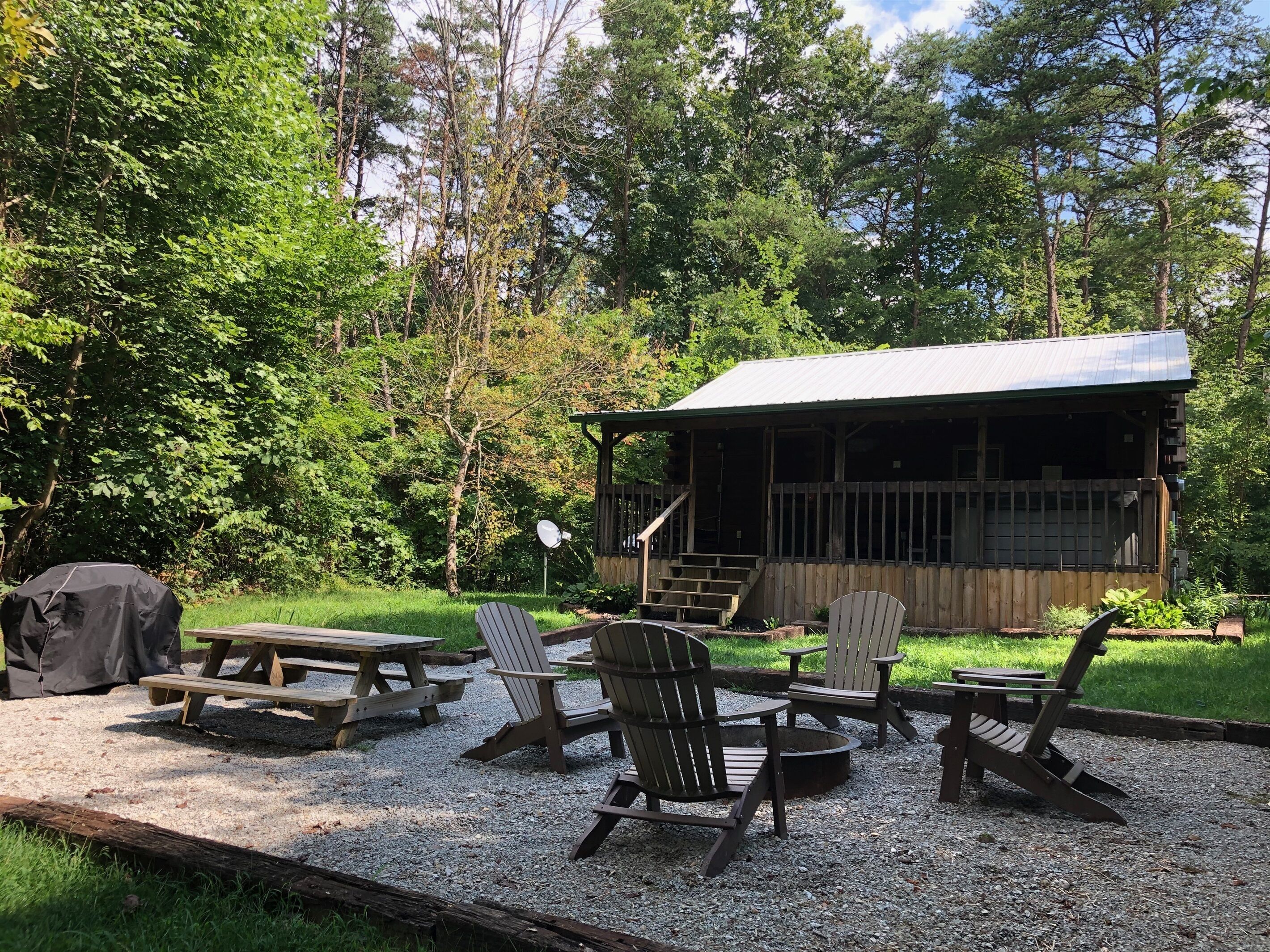 A rustic cabin with a covered porch, picnic tables, and Adirondack chairs in a wooded area.